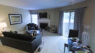 This single-floor space centers around a well-appointed living area that balances comfort and modern style. The room features a white-mantled fireplace serving as a natural focal point, topped with a wall-mounted flat-screen television. Soft, neutral-toned carpeting runs throughout, complementing the light gray walls and creating a warm, cohesive atmosphere. Natural light flows generously into the space through two windows and a sliding glass door, the latter framed by bold black-and-white geometric patterned curtains that add a striking visual element. The sliding door opens to an outdoor balcony or patio area, offering views of a lush green landscape beyond. The living area is furnished with dark leather seating, including a sofa and armchair, arranged around a square coffee table. A glass-topped dining table with dark chairs is positioned near the sliding door, creating a defined dining area within the open layout. Decorative accessories throughout maintain a consistent monochromatic black-and-white theme, lending the space a cohesive and intentional design aesthetic.