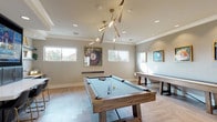 This well-appointed single-floor amenity space is designed with entertainment and social interaction in mind. The main game room serves as the centerpiece, showcasing a full-size pool table with a light blue felt surface, complemented by a shuffleboard table along the perimeter wall. The room features striking herringbone-patterned light wood flooring throughout, which lends a refined yet approachable character to the space. A sculptural, branch-inspired gold chandelier hangs above, providing both ambient and task lighting alongside recessed ceiling fixtures. Natural light filters in through multiple horizontal windows positioned along the upper portion of the walls, offering views of the surrounding landscape. Billiard-themed artwork in gold frames adds a cohesive decorative touch throughout. Along one wall, a built-in wet bar and lounge station features dark cabinetry, a large flat-screen TV, open floating shelves, a wine refrigerator, a dishwasher, a sink, and a bar-height countertop with seating. A small transitional area with additional seating and a frosted-glass door leads to an adjacent media room, providing a quieter retreat. The overall ambiance is modern and sophisticated, balancing functionality with an upscale aesthetic suited for communal gatherings.