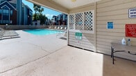 The pool facility at 3131 Cavalry Dr in Raleigh, North Carolina is a single-level, residents-only outdoor amenity space designed for relaxation and recreation. The area is accessed through a covered entryway featuring white lap siding walls, crown molding details, and a dark-framed glass door. The entry vestibule includes an emergency phone station and posted pool safety guidelines, ensuring resident awareness and security.

Beyond the entry, a gated pool pass checkpoint with white lattice panels and a wooden fence leads to the open-air pool deck. The swimming pool features clear blue water surrounded by a generous concrete surround, offering ample space for lounging. A row of reclining pool chairs lines the perimeter, ideal for sunbathing. A charcoal grill station provides an outdoor cooking option, while a patio table shaded by a turquoise umbrella creates a comfortable gathering spot.

The covered pavilion area adjacent to the pool offers a shaded retreat, framed by decorative white lattice panels that allow natural light and breezes to filter through. The surrounding wooden privacy fence encloses the space, with views of mature trees and the broader residential community beyond, contributing to a pleasant and community-oriented outdoor atmosphere.