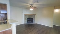 This single-floor space offers a straightforward and functional layout centered around a welcoming living area. The focal point of the room is a corner fireplace clad in natural stone with a wood mantel, adding warmth and character to the space. Dark hardwood-style flooring runs throughout, providing a rich, grounding contrast to the neutral, light-toned walls. A ceiling fan with an integrated light fixture is centrally positioned, ensuring both air circulation and ambient lighting. The textured popcorn ceiling adds a classic residential touch. A partial countertop or bar-height surface is visible at the edge of the frame, suggesting an open connection to an adjacent kitchen or dining area. The open layout of the living space allows for flexible furniture arrangement. Overall, the space presents a comfortable and practical interior with functional features suited for everyday living.