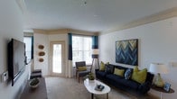 This single-floor residence is thoughtfully designed to offer a comfortable and contemporary living experience. The living room serves as the heart of the space, showcasing a neutral color palette accented with bold teal and chartreuse tones that lend a vibrant, modern energy to the interior. Crown molding frames the ceiling, adding an elegant architectural detail, while a textured ceiling finish provides subtle character throughout. Large windows dressed with two-tone teal and navy curtains flood the room with natural light, creating a warm and inviting atmosphere. A private exterior door offers convenient access to an outdoor area, further enhancing the sense of openness. The living area is generously proportioned, comfortably accommodating a full-sized sofa, accent chair, and a round coffee table, making it well-suited for both relaxing and entertaining. Wall art featuring a bold geometric chevron design adds a striking visual focal point, complementing the contemporary furnishings. Plush carpeted flooring runs throughout, adding warmth and comfort underfoot. Overall, the space presents a well-balanced blend of stylish design and functional living, with carefully curated décor and abundant natural light defining its welcoming ambiance.