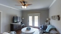 This single-floor space presents a thoughtfully designed living area that balances comfort and classic style. The living room serves as the focal point of the home, showcasing rich hardwood flooring that grounds the space with warmth and durability. Neutral gray walls create a versatile backdrop that complements the room's furnishings and décor.

Crown molding lines the ceiling, adding a refined architectural detail that elevates the overall aesthetic. A ceiling fan with a light kit provides both practical air circulation and ambient lighting. The room is anchored by a media console with ample drawer storage, positioned across from a comfortable seating arrangement accented with navy and white patterned throw pillows.

A round white coffee table sits at the center of the living area, atop a light-toned area rug that defines the space. The room benefits from abundant natural light, which streams in through a set of white-trimmed French doors. These doors not only enhance the brightness of the interior but also provide direct access to an outdoor area, offering a seamless connection between indoor and outdoor living. Accent lighting from coordinating table and floor lamps adds layered illumination throughout. Overall, the space presents a clean, classic design with functional and inviting details.