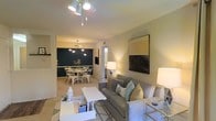 This well-appointed single-floor residence offers a seamless blend of comfort and contemporary style. The open-concept layout connects the living and dining areas, creating a spacious, airy feel ideal for both relaxing and entertaining. The living area is anchored by a plush gray sofa accented with teal and patterned throw pillows, complemented by a clean white coffee table and layered area rug. A bold mixed-media wall art piece adds a striking focal point to the room, while warm ambient lighting from floor lamps creates an inviting atmosphere. Track lighting on the ceiling provides additional illumination and a modern touch overhead. The dining area is defined by a deep teal accent wall that adds dramatic contrast to the otherwise neutral color palette. A round dining table surrounded by sleek metal chairs sits beneath a mid-century modern chandelier, lending the space a curated, designer feel. Natural light filters in through a nearby window, brightening the transition between the two zones. Soft carpeting runs throughout the main living areas, adding warmth underfoot. The overall ambiance is cohesive and thoughtfully styled, with a neutral base palette punctuated by bold accents, making this a comfortable and visually engaging space.