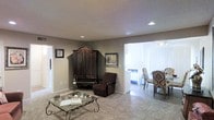 This single-floor residence offers a comfortable and well-appointed layout designed for easy living. The living area serves as the heart of the home, featuring soft wall-to-wall carpeting underfoot and a neutral, warm color palette on the walls that creates an inviting atmosphere. Recessed lighting in the ceiling provides consistent, even illumination throughout the space. A decorative glass-top coffee table with ornate wrought-iron legs anchors the seating area, complemented by a plush recliner chair and a striking dark wood armoire that adds a classic, traditional character to the room. Framed artwork adorns the walls, contributing to the space's homey aesthetic. An open passageway connects the living area to a bright dining space, which benefits from abundant natural light filtering through large vertical blinds. The dining area features hardwood-style flooring, creating a pleasant contrast to the carpeted living room. The transition between the two areas is seamless, enhancing the open and flowing feel of the floor plan. Additional doorways suggest further rooms beyond the main living space, completing this well-organized and warmly decorated single-story home.