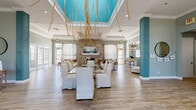 Located at 200 Village Blvd in Davenport, Florida, this single-story community clubhouse offers an inviting and well-appointed gathering space designed for both social events and everyday relaxation. The centerpiece of the facility is a spacious great room distinguished by soaring tray ceilings with a vibrant teal inset, elegant crown molding, and wood-look flooring that flows throughout. A dramatic floor-to-ceiling stone fireplace anchors the lounge area, flanked by plush seating and a rustic live-edge coffee table, creating a warm and comfortable atmosphere. A glass-top dining table with slipcovered chairs provides a dedicated space for communal dining and activities. The entryway features teal decorative columns, a console table, and easy access to French door exits that bathe the space in natural light. An arched transom window above the main entrance further enhances the bright, airy feel. A fully equipped kitchen with dark cabinetry, granite countertops, stainless steel appliances, and a coffee station supports catering and resident use. A separate sitting area with French doors and wicker accent chairs offers a quieter retreat. Large windows dressed with sheer curtains ensure the entire space remains light-filled, while the open layout provides versatility for a wide range of community functions.