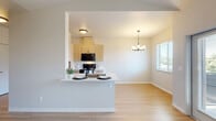 This thoughtfully laid-out one-bedroom, one-bathroom residence offers a clean, modern aesthetic across its single floor. Light wood-style flooring runs continuously throughout, creating a cohesive and airy feel.

The galley-style kitchen is a standout feature, lined on both sides with light wood cabinetry extending to the ceiling for ample storage. Black appliances — including a refrigerator, range, microwave, and dishwasher — provide a bold contrast against the white countertops. A peninsula with a breakfast bar extends into the open living and dining area, which is illuminated by a pendant chandelier and a large window that draws in natural light.

The bedroom is a comfortable, well-proportioned space featuring vaulted ceilings, a ceiling fan, a window with an outdoor view, and a built-in closet with a sliding door and full-length hanging rod. A second window-lit room offers flexibility as an additional bedroom or dedicated workspace.

The bathroom features a wood-toned vanity with a white countertop, a large mirror, and a shower/tub combination with a curtain rod. White walls and light flooring keep the space feeling clean and open. A wall-mounted HVAC unit in the living area ensures year-round comfort throughout this well-appointed residence.