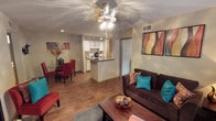 This single-floor space is designed around an open-concept floor plan that seamlessly connects the living, dining, and kitchen areas into one cohesive, functional environment. The living area is anchored by warm hardwood-style flooring that extends throughout the unit, creating a consistent and inviting aesthetic. A ceiling fan with integrated lighting serves as a central fixture overhead, complementing recessed lighting that provides additional illumination across the space.

The kitchen features white cabinetry, a dark countertop peninsula that doubles as a breakfast bar, and stainless steel appliances, including a full-size refrigerator. Track lighting above the kitchen adds focused task lighting to the area. The peninsula serves as a natural divider between the kitchen and the main living space without disrupting the open feel.

Adjacent to the living room, the dining area accommodates a small table and chairs, making it well-suited for casual meals. Colorful wall art adds personality throughout, with vibrant accents complementing the neutral beige walls. The overall ambiance is warm, practical, and well-lit, making efficient use of the available space.
