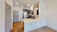 Located at 207 Gresham Ln in Mooresville, NC, this modern single-floor residence showcases a clean, contemporary design throughout. Warm wood-look flooring flows seamlessly across all rooms, creating a cohesive and inviting atmosphere.

The kitchen is a standout feature, outfitted with stainless steel appliances including a refrigerator, range, and microwave. Dark cabinetry with matte black hardware contrasts sharply against crisp white quartz countertops, while a subway tile backsplash adds texture. A peninsula with pendant lighting and an undermount sink with a matte black faucet provides both additional workspace and a natural gathering point that connects to the main living area. A built-in wine rack adds a refined touch.

The open living area benefits from recessed lighting and abundant natural light, offering a versatile and airy space. The bedroom is well-proportioned, featuring a ceiling fan with integrated lighting and a window that invites natural light and outdoor views.

The bathroom is finished to a high standard, featuring a dark vanity with a white countertop, a large mirror with modern lighting, and a soaking tub with a tiled surround and matte black fixtures. The overall design palette — white walls, dark accents, and warm flooring — gives this residence a polished, move-in-ready appeal.