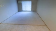 This single-floor space presents a straightforward and functional layout highlighted by clean, neutral aesthetics throughout. The room visible in the image features wall-to-wall carpeting in a light, neutral tone that contributes to a calm and understated atmosphere. The walls are painted in a soft, muted shade that complements the flooring and maintains a cohesive, understated palette.

A large window dressed with vertical blinds serves as the room's focal point, allowing natural light to filter in and brighten the interior. The transition between the carpeted area and an adjacent hard-surface flooring section is visible at the foreground, suggesting a connected space with varied flooring materials — a practical design detail that helps define different functional zones.

The baseboard trim runs consistently along the walls, and electrical outlets are positioned at standard heights, indicating a move-in-ready condition. The overall ambiance is clean, quiet, and neutral, offering a blank canvas that can be adapted to a range of uses. The space's simplicity and light-filled quality make it a practical and welcoming environment.