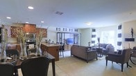 This single-floor residence features an open-concept layout that unifies the kitchen, dining, and living areas into one expansive, flowing space. The kitchen is equipped with stainless steel appliances, including a range and microwave, and features warm wood cabinetry paired with a granite-topped island that doubles as a breakfast bar with seating. Recessed lighting illuminates the kitchen area, complementing the neutral-toned walls throughout.

The dining area transitions naturally from the kitchen, offering an intimate setting for meals. Beyond it, the living room is anchored by a stylish open shelving unit with a media console, displaying decorative accents and housing a flat-screen television. The seating arrangement includes a plush sofa and coordinating chairs in neutral tones, placed atop a textured area rug that defines the space.

Natural light filters in through a window dressed with bold striped curtains, adding a graphic design element to the room. Decorative touches, including tall branches in vases and accent pillows, contribute to the polished, cohesive aesthetic. The light-colored flooring and neutral palette create a bright, welcoming ambiance throughout.