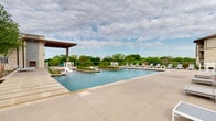 This single-story amenity facility is defined by its striking limestone exterior, which lends a refined, Hill Country-inspired aesthetic throughout. The architecture integrates natural stone masonry, warm wood-planked ceilings, and bold black-framed glass windows and doors, creating a cohesive and upscale visual identity.

The outdoor spaces are the centerpiece of the facility. A resort-style pool area stretches generously across the grounds, featuring a lap pool accessible via a stone-flanked entry corridor with handrails, alongside a larger leisure pool framed by manicured plantings and rows of contemporary white lounge chairs. A covered cabana with a modern flat roof provides shaded seating and overlooks the pool deck, which is finished in large-format concrete pavers. The open sky and surrounding treeline create a serene, nature-connected ambiance.

Covered verandas wrap portions of the building's perimeter, offering shaded outdoor seating beneath wood-paneled ceilings supported by chunky limestone columns. These walkways connect multiple entry points, each featuring glass double doors that allow natural light to flow freely into interior spaces.

An outdoor kitchen area is built into one of the stone walls, equipped with a stainless steel grill, sink, counter space, and a wall-mounted television — ideal for outdoor gatherings. A controlled-access entry with glass gate panels and elegant pendant lighting marks a formal point of entry into the complex.