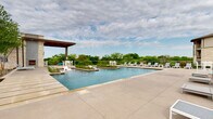 This single-story amenity facility is defined by its striking limestone exterior, which lends a refined, Hill Country-inspired aesthetic throughout. The architecture integrates natural stone masonry, warm wood-planked ceilings, and bold black-framed glass windows and doors, creating a cohesive and upscale visual identity.

The outdoor spaces are the centerpiece of the facility. A resort-style pool area stretches generously across the grounds, featuring a lap pool accessible via a stone-flanked entry corridor with handrails, alongside a larger leisure pool framed by manicured plantings and rows of contemporary white lounge chairs. A covered cabana with a modern flat roof provides shaded seating and overlooks the pool deck, which is finished in large-format concrete pavers. The open sky and surrounding treeline create a serene, nature-connected ambiance.

Covered verandas wrap portions of the building's perimeter, offering shaded outdoor seating beneath wood-paneled ceilings supported by chunky limestone columns. These walkways connect multiple entry points, each featuring glass double doors that allow natural light to flow freely into interior spaces.

An outdoor kitchen area is built into one of the stone walls, equipped with a stainless steel grill, sink, counter space, and a wall-mounted television — ideal for outdoor gatherings. A controlled-access entry with glass gate panels and elegant pendant lighting marks a formal point of entry into the complex.