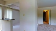 This single-floor space presents a clean, functional layout characterized by its neutral color palette and open interior design. The walls are finished in soft, light tones that contribute to a bright and airy ambiance throughout. A prominent structural column serves as a defining architectural element, subtly dividing the space while maintaining an open feel between the connected areas. The flooring transitions from light-tiled surfaces in one section to a contrasting material in an adjoining area, adding visual interest and helping to define distinct zones within the open plan. A glimpse of a countertop edge hints at a kitchen or utility area to one side, while the open passageways suggest a flowing connection between the living spaces. The layout is straightforward and accessible, with standard wall outlets and switches positioned throughout. Natural light filters into the space, enhancing the brightness of the pale walls and contributing to the overall open, welcoming atmosphere. The simplicity of the interior design provides a versatile canvas, well-suited for a variety of uses and furnishing styles.