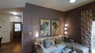 This thoughtfully designed single-floor space makes a strong impression through its carefully curated interior aesthetic. The living area serves as the heart of the space, anchored by deep mauve-toned walls that lend a warm, sophisticated atmosphere. A generously sized sectional sofa in soft neutral upholstery provides ample seating and is dressed with an array of decorative pillows in muted greens, golds, and grays, adding layered texture and color. Two striking large-format equestrian paintings hang prominently on the feature wall, becoming an immediate focal point and lending an artistic character to the room. Lighting plays a key role in defining the ambiance — a sculptural multi-globe floor lamp and a delicate crystal-accented wall sconce cast soft, decorative light that dances across the walls, adding depth and warmth to the space. A rustic wooden side table grounds the seating area with a natural contrast to the refined décor. The flooring features warm wood tones that complement the overall color palette. The space is further elevated by clean white ceilings that balance the richness of the walls, creating a polished and cohesive environment that feels both inviting and refined.