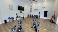 This single-floor fitness studio offers a clean, well-organized space designed to support a variety of workout routines. The room features light blue walls and warm wood-tone flooring, creating a bright and welcoming atmosphere. A large wall-mounted flat-screen TV serves as a focal point at the front of the room, accompanied by a soundbar, making it ideal for guided workout sessions or virtual classes. Several spin bikes are positioned toward the front, providing a dedicated cycling area. Along the back wall, a neatly organized dumbbell rack holds a full range of free weights, while additional equipment racks store medicine balls, foam rollers, and resistance tools. White storage shelving units flank the TV, offering convenient organization for smaller accessories. Crown molding adds a polished architectural touch, while recessed lighting and a ceiling fan ensure the space remains comfortable during intense workouts. A decorative "RELAX" sign and framed artwork contribute a welcoming, resort-like ambiance. The open floor plan allows for flexible use of the space beyond the fixed equipment, accommodating stretching, bodyweight exercises, or small group training sessions.