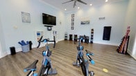 This single-floor fitness studio offers a clean, well-organized space designed to support a variety of workout routines. The room features light blue walls and warm wood-tone flooring, creating a bright and welcoming atmosphere. A large wall-mounted flat-screen TV serves as a focal point at the front of the room, accompanied by a soundbar, making it ideal for guided workout sessions or virtual classes. Several spin bikes are positioned toward the front, providing a dedicated cycling area. Along the back wall, a neatly organized dumbbell rack holds a full range of free weights, while additional equipment racks store medicine balls, foam rollers, and resistance tools. White storage shelving units flank the TV, offering convenient organization for smaller accessories. Crown molding adds a polished architectural touch, while recessed lighting and a ceiling fan ensure the space remains comfortable during intense workouts. A decorative "RELAX" sign and framed artwork contribute a welcoming, resort-like ambiance. The open floor plan allows for flexible use of the space beyond the fixed equipment, accommodating stretching, bodyweight exercises, or small group training sessions.
