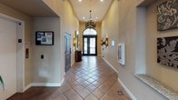 This lobby showcases a refined Spanish Colonial aesthetic, offering a welcoming and elegant first impression. The corridor-style layout is anchored by striking terracotta tile flooring set in a diagonal pattern, accented with decorative mosaic tile inlays at select intervals. The soaring vaulted and barrel-arched ceilings add a sense of grandeur, drawing the eye upward and emphasizing the generous vertical space throughout.

The grand double entry doors serve as a stunning focal point, featuring ornate wrought iron scrollwork and a dramatic arched transom window that floods the space with natural light. Decorative wrought iron wall sconces line the corridor, casting a warm, ambient glow that complements the soft, neutral wall tones.

Overhead, an intricately detailed pendant chandelier with a wrought iron frame adds a sophisticated centerpiece to the space. A curved, dark wood reception console sits near the entry, providing both function and style. Architectural niches along the walls offer display space, and artwork adds curated visual interest throughout. A lush potted plant near the entry doors introduces a natural element, softening the overall composition. The lobby presents a cohesive, upscale environment that blends traditional craftsmanship with timeless design details.