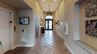 This lobby showcases a refined Spanish Colonial aesthetic, offering a welcoming and elegant first impression. The corridor-style layout is anchored by striking terracotta tile flooring set in a diagonal pattern, accented with decorative mosaic tile inlays at select intervals. The soaring vaulted and barrel-arched ceilings add a sense of grandeur, drawing the eye upward and emphasizing the generous vertical space throughout.

The grand double entry doors serve as a stunning focal point, featuring ornate wrought iron scrollwork and a dramatic arched transom window that floods the space with natural light. Decorative wrought iron wall sconces line the corridor, casting a warm, ambient glow that complements the soft, neutral wall tones.

Overhead, an intricately detailed pendant chandelier with a wrought iron frame adds a sophisticated centerpiece to the space. A curved, dark wood reception console sits near the entry, providing both function and style. Architectural niches along the walls offer display space, and artwork adds curated visual interest throughout. A lush potted plant near the entry doors introduces a natural element, softening the overall composition. The lobby presents a cohesive, upscale environment that blends traditional craftsmanship with timeless design details.