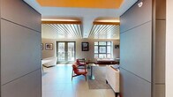 Located at 42 Broad St W in Mount Vernon, NY, this thoughtfully designed amenity space is housed within a striking multi-story brick and glass building. The interior showcases a warm, contemporary aesthetic anchored by distinctive slatted wood ceilings that run throughout, lending a sense of texture and architectural character to the entire floor.

The open living area serves as the centerpiece of the space, featuring a curated seating arrangement with mid-century modern chairs in earthy terracotta tones, a comfortable sofa, and a mounted flat-screen display set against a richly colored accent wall. Framed artwork adorns the walls, contributing to an inviting, gallery-like atmosphere. Large windows flood the space with natural light and offer views of the surrounding streetscape.

Adjacent to the lounge, a sleek kitchen island with a light wood finish and integrated appliances provides a functional hub for gatherings, complemented by large-format tile flooring that extends throughout. A defined dining area with a communal table and modern pendant-style chairs sits alongside a textured feature wall, creating a distinct zone for shared meals or meetings.

Outdoors, a well-appointed courtyard grilling station equipped with professional-grade stainless steel grills offers an ideal setting for al fresco entertaining, framed by the building's contemporary brick and panel facade.