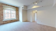 This single-floor residence offers a compelling blend of historic architectural character and contemporary finishes. The open living area immediately draws attention with its striking exposed brick accent wall, soaring ceilings, and rail track lighting that highlights the generous proportions of the space. Soft wall-to-wall carpeting runs throughout the living and bedroom areas, adding warmth underfoot.

Large windows frame views of classic urban architecture, allowing natural light to fill the interior. A dedicated bedroom features its own exposed brick wall framed around an arched window surround, lending it a distinctive loft-like character, along with a spacious closet fitted with hanging rods for ample storage.

An additional room with a broad window overlooking neighboring historic buildings offers flexibility as a second bedroom, office, or den. The kitchen is outfitted with dark cabinetry, granite-style countertops, and a full suite of black appliances including a range, microwave, and refrigerator, all set over easy-clean tile flooring.

Two bathrooms serve the residence: one featuring a full soaking tub with tiled surround, and a second with a stand-alone shower, dark vanity cabinetry, and a wide mirror. Together, these spaces create a functional and characterful urban home rich in architectural detail.