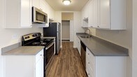 This completely remodeled 1-bedroom, 1-bathroom unit showcases thoughtful updates throughout its single-floor layout. The open living area features plush neutral carpeting, clean white crown molding, and a ceiling fan, creating a comfortable and well-finished environment. A wall-mounted HVAC vent ensures year-round climate control. The living space flows into a secondary carpeted room highlighted by a bold terracotta accent wall and a sliding glass door fitted with vertical blinds, offering views of the outdoors and access to a balcony.

The galley-style kitchen is efficiently designed with white shaker cabinetry, gray laminate countertops, wood-look vinyl plank flooring, and a full suite of stainless steel appliances including a refrigerator, range, microwave, and dishwasher. A closet visible at the end of the kitchen provides additional storage convenience.

The bedroom continues the warm, carpeted aesthetic and also features a terracotta accent wall alongside a large sliding window with vertical blinds, allowing ample natural light. The remodeled bathroom is appointed with wood-look vinyl flooring, white shaker vanity cabinetry, a dark granite countertop, an undermount sink with brushed nickel fixtures, and a tub-shower combination enclosed by sliding glass doors.