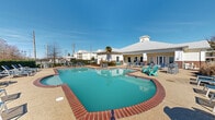 Located at 7801 Scenic Hwy in Baton Rouge, Louisiana, this outdoor amenity area centers around a generously sized swimming pool with a distinctive curved design and classic brick coping. The pool's clear turquoise water is complemented by a wide, aggregate-finished sun deck that wraps around the perimeter, providing ample space for lounging. The deck is well-furnished with an assortment of blue and teal lounge chairs, Adirondack chairs, and bar-height seating, offering a variety of relaxation options for residents. Stainless steel entry rails provide convenient pool access.

The single-story clubhouse building anchors the space with a traditional Southern architectural style, featuring white siding, brick accents, a hipped roof with light-colored shingles, and French-style doors that open directly onto the pool deck. A palm tree near the building adds a subtropical touch that complements the Louisiana setting. Manicured hedgerows and landscaped beds line portions of the perimeter, providing a degree of privacy and a lush green backdrop. A wrought iron fence encloses the area, ensuring security while maintaining open sightlines. The surrounding residential buildings are visible beyond the enclosure, situating this amenity space as a central gathering hub within the broader community.