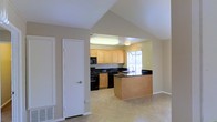 This single-story residence showcases a thoughtfully designed open-concept layout that promotes a seamless flow between living and kitchen spaces. The living area features striking vaulted ceilings that create a sense of openness and airiness, with clean neutral wall tones complementing the light-toned flooring throughout. A wide archway connects the living space to the kitchen, creating an inviting and functional transition between the two areas. The kitchen is well-equipped and features light maple cabinetry that provides ample storage, contrasted elegantly by dark granite-style countertops. A peninsula island serves as both a prep space and a casual gathering point. Black appliances, including a range and microwave, add a sleek, modern touch to the kitchen's aesthetic. A large window above the sink allows natural light to pour into the kitchen, brightening the space and offering a pleasant outlook. The kitchen also benefits from dedicated overhead lighting, ensuring the workspace remains well-lit at all times. Light tile flooring runs throughout the kitchen and into the living area, providing a cohesive and easy-to-maintain surface. A flush interior door with clean white trim adds a crisp, finished look to the overall design. The space presents a practical and comfortable single-floor living environment.