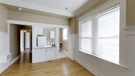 This single-floor space showcases a blend of classic craftsmanship and functional design. The main living area is distinguished by warm hardwood floors and elegant crown molding that frames the ceiling, lending the space a timeless character. Neutral taupe walls create a calm, versatile backdrop throughout. A large window fitted with horizontal blinds floods the living area with natural light, enhancing the open, airy feel of the room. A decorative chair rail runs along the lower portion of the walls, adding an additional layer of traditional architectural detail.

The kitchen is accessible through a wide, framed opening that maintains a visual connection between the two spaces. It features white cabinetry, a light-toned countertop, and a white refrigerator, all contributing to a clean and cohesive aesthetic. A window above the kitchen area provides additional natural light. An adjacent nook or hallway area offers a small built-in desk or ledge, adding a practical element to the layout. White trim and baseboards are consistent throughout, tying the spaces together cohesively and reinforcing the unit's classic, well-maintained character.
