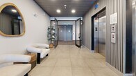 Located at 46120 Thomas Rd in Chilliwack, BC, this building lobby offers a polished and welcoming first impression. The entrance is defined by floor-to-ceiling glass walls framed in sleek black steel, flooding the space with abundant natural light and providing clear views to the surrounding outdoor environment. Large-format neutral-toned floor tiles extend throughout, lending a clean, contemporary aesthetic. The double-height ceiling in the main entrance area creates a dramatic sense of openness, and is adorned with a striking sculptural pendant light fixture that serves as a focal point. Accessibility is a clear priority, with automatic doors and clearly marked activation panels. Beyond the entrance vestibule, the interior corridor transitions to a more intimate scale, featuring a dark slatted ceiling, recessed lighting, and textured wall paneling that adds warmth and depth. This inner hallway includes a comfortable seating area with a white upholstered bench, a decorative indoor plant, and direct access to an elevator. Additional functional elements include a building intercom system, fire alarm panel, and mailbox area, making this lobby a comprehensive and well-appointed building entry point.