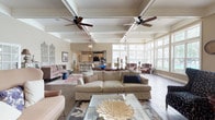 Located at 5303 Wilcox Rd in Dublin, Ohio, this single-floor community space offers a refined and welcoming atmosphere designed for both relaxation and socializing. The interior is defined by soaring coffered ceilings adorned with ceiling fans and pendant lantern-style light fixtures, creating a warm and sophisticated ambiance throughout.

The expansive main gathering area is anchored by large floor-to-ceiling windows that flood the space with natural light and offer views of the surrounding outdoors. Elegant herringbone-patterned wood-look tile flooring runs throughout, adding a timeless, classic character to each room. A standout feature is the fireplace flanked by built-in shelving and cabinetry, set against a rich navy blue accent wall, providing a cozy and stylish focal point.

Comfortable and thoughtfully curated seating arrangements — including tufted sofas, wingback chairs, and accent tables — are distributed across multiple lounge zones, allowing for both intimate conversations and larger gatherings. Decorative wall art and carefully chosen accessories contribute to the space's upscale, residential feel.

The full kitchen is equipped with white cabinetry, granite countertops, black appliances, and a generously sized island with bar seating, making it ideal for catered events or community gatherings. French doors provide seamless access to exterior areas, further enhancing the open, airy feel of this well-appointed community clubhouse.
