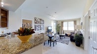 This single-floor, two-bedroom, two-bathroom residence features an open-concept design that seamlessly integrates the kitchen, dining, and living areas into one cohesive, flowing space. The kitchen is appointed with granite countertops featuring a curved breakfast bar overhang, providing both a functional workspace and a casual dining option. The countertops display rich, warm tones with natural veining, adding a touch of elegance to the space.

The open dining area accommodates a full dining table and chairs, positioned conveniently between the kitchen and living room, making it ideal for both everyday meals and entertaining. The living room is generously proportioned, featuring neutral-toned carpeting, and is brightened by large windows dressed with curtains that allow ample natural light to filter in. Track lighting along the ceiling provides additional illumination throughout the main living area.

The overall aesthetic is modern and inviting, with neutral wall tones complemented by decorative wall art and lush indoor plants that add warmth and character. The layout promotes an airy, connected feel throughout the main living space.