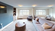 This well-appointed single-floor space is designed to foster comfort and social interaction through a series of thoughtfully arranged seating areas. The layout flows between two distinct zones: a carpeted lounge anchored by a plush sofa, accent chairs, and a distinctive round wooden coffee table, and an open hardwood-floored gathering area furnished with an inviting arrangement of upholstered chairs and small side tables.

The carpeted lounge area is enhanced by a bold navy blue accent wall that frames a wall-mounted flat-screen television set within built-in shelving and cabinetry, providing both display and storage functionality. A small kitchenette alcove with dark cabinetry is conveniently situated nearby. Multiple windows along the perimeter allow generous natural light to fill the space, offering views of lush greenery outdoors.

The open hardwood-floored seating area features warm, neutral tones throughout, with light wood plank flooring that adds warmth and continuity. A large framed abstract artwork in deep blues and golds serves as a striking focal point on one wall, complemented by the presence of tall indoor plants that bring a touch of nature inside. Flush-mount ceiling fixtures provide consistent ambient lighting throughout. Together, these areas create a versatile, welcoming environment suited for relaxed gatherings or quiet conversation.