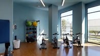 This well-appointed fitness center offers a clean, modern environment designed to support a wide range of workout routines. The space is finished with warm hardwood-style flooring that contrasts beautifully against the cool blue-gray walls, creating an energizing yet calming atmosphere. Overhead, linear LED lighting complements the abundant natural light that streams in through large floor-to-ceiling windows, offering views of the outdoors and filling the room with a bright, open feel. The cycling area is equipped with a row of stationary spin bikes, thoughtfully arranged to allow ample space between each station. On the opposite side of the room, a well-stocked equipment rack holds an assortment of functional training tools, including stability balls, medicine balls, kettlebells, resistance bands, and TRX suspension straps, catering to a variety of fitness levels and training styles. Structural columns are integrated seamlessly into the layout without disrupting the flow of the space. The overall design is both practical and inviting, striking a balance between a professional gym environment and an accessible community fitness facility. The single-floor layout ensures effortless navigation throughout the space.