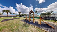 This outdoor amenity area is a thoughtfully designed pet-friendly and recreational space situated within a residential community surrounded by lush palm trees and mature shade trees. The grounds are enclosed by a black wrought iron fence, providing a secure and well-defined perimeter throughout the entire area.

At the heart of the space is a dedicated dog park featuring purpose-built canine agility and play equipment, including a multi-level wooden play structure adorned with paw print details, a tunnel, a climbing ramp, and a slide. The agility area is set on a rubberized mulch surface for safety and comfort. Adjacent to the dog play zone, a separate agility course offers additional obstacles for pets to explore.

Beyond the dog park, the open green lawn provides generous space for outdoor recreation. A set of blue Adirondack chairs invites residents to relax in the fresh air, while a cornhole board setup encourages casual outdoor games. A sandbox adds an additional recreational feature to the grounds.

The well-maintained grass lawn, dotted with tall palm trees, creates a vibrant, tropical ambiance that is sunny and inviting. Residential buildings with balconies border the space, providing a sense of community and easy access to the amenities. The area benefits from abundant natural light and open sky views, making it an enjoyable outdoor destination for residents and their pets alike.