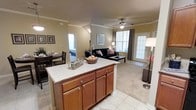 This single-floor residence offers a thoughtfully designed open-concept layout that brings together the kitchen, dining, and living spaces in a cohesive and functional arrangement. The kitchen is anchored by a central island with a tiled countertop and rich warm-toned cabinetry, providing ample storage and prep space. Tile flooring runs through the kitchen and transitions smoothly into the surrounding areas, lending a clean and consistent look throughout.

The dining area sits just off the kitchen, comfortably accommodating a full dining set and benefiting from natural light filtering in from nearby windows. The living room is spacious and inviting, featuring plush carpeting, crown molding, and a ceiling fan that adds both style and comfort. A large window and glass-paneled door flood the space with natural light and offer a connection to the outdoors.

The warm, neutral color palette throughout creates a welcoming ambiance, while classic architectural details such as crown molding and column accents add a touch of elegance. Additional rooms are visible beyond the main living area, suggesting a well-rounded floor plan designed for comfortable everyday living. Overall, this residence balances practicality with a warm, homey aesthetic.