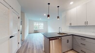 This single-floor residence showcases a clean, contemporary design with a cohesive aesthetic carried throughout every room. The open-concept layout connects the kitchen and living area seamlessly, creating a bright and functional shared space anchored by light-toned luxury vinyl plank flooring. The kitchen is well-equipped with stainless steel appliances, including a refrigerator, electric range, and built-in microwave. A central island with a dark countertop and an undermount sink serves as both a prep space and a natural divider, illuminated by two sleek pendant lights overhead. White upper cabinetry and gray lower cabinets provide ample storage, complemented by a classic subway tile backsplash. The living area benefits from natural light through a large window with views of surrounding greenery. The bedrooms are bright and well-sized, each featuring large windows framing lush outdoor tree views, light wood-tone flooring, and individual climate control units. The two bathrooms are finished with large-format neutral tiles, white vanities, and a glass-enclosed walk-in shower in the primary bath, along with accessibility-friendly features such as grab bars. Recessed lighting throughout ensures a well-lit, welcoming ambiance across the entire space.