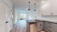 This single-floor residence showcases a clean, contemporary design with a cohesive aesthetic carried throughout every room. The open-concept layout connects the kitchen and living area seamlessly, creating a bright and functional shared space anchored by light-toned luxury vinyl plank flooring. The kitchen is well-equipped with stainless steel appliances, including a refrigerator, electric range, and built-in microwave. A central island with a dark countertop and an undermount sink serves as both a prep space and a natural divider, illuminated by two sleek pendant lights overhead. White upper cabinetry and gray lower cabinets provide ample storage, complemented by a classic subway tile backsplash. The living area benefits from natural light through a large window with views of surrounding greenery. The bedrooms are bright and well-sized, each featuring large windows framing lush outdoor tree views, light wood-tone flooring, and individual climate control units. The two bathrooms are finished with large-format neutral tiles, white vanities, and a glass-enclosed walk-in shower in the primary bath, along with accessibility-friendly features such as grab bars. Recessed lighting throughout ensures a well-lit, welcoming ambiance across the entire space.