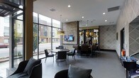This stylish single-floor communal space blends contemporary design with warm, upscale finishes to create an inviting and functional environment. Floor-to-ceiling windows along one wall flood the interior with natural light while offering open views of the surrounding streetscape, giving the space an airy, connected feel.

The layout is thoughtfully organized into distinct seating and gathering zones. A casual lounge area near the windows features plush, dark upholstered seating arranged for relaxed conversation. Adjacent to this, a round dining table with sleek chairs provides an intimate spot for smaller gatherings.

A striking stacked-stone accent wall serves as a dramatic focal point, anchoring a wall-mounted flat-screen television and adding a rich, textural contrast to the otherwise clean, modern aesthetic. Nearby, a bar-height dining area with leather-toned chairs offers a more social setting, illuminated by a cluster of bold, gold globe pendant lights that add a touch of glamour and warmth.

The flooring throughout consists of large-format grey tile, contributing to the space's polished and cohesive look. Recessed lighting complements the decorative pendant fixtures, ensuring the space is well-lit at all hours. The overall ambiance is modern and refined, ideal for socializing, dining, or unwinding in a well-appointed setting.