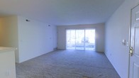 This single-floor space features an open living area finished with wall-to-wall carpeting in a neutral gray tone, contributing to a clean and understated aesthetic. The room is bright and airy, thanks largely to a full-width sliding glass door that serves as the focal point of the main living area. The glass door opens to an outdoor balcony or patio, allowing generous natural light to flood the interior while providing a direct connection to the exterior. Vertical blinds frame the sliding door, offering flexibility in light control and privacy. The walls are painted in a crisp white throughout, complementing the light tones of the carpet and reinforcing the open, uncluttered feel of the space. A textured white ceiling adds subtle depth overhead, while a standard interior door on one side of the room leads to an additional area of the unit. Ceiling vents and floor-level HVAC registers indicate a central climate control system. The overall layout is straightforward and functional, offering a blank canvas well-suited for a variety of furniture arrangements and personal styles.