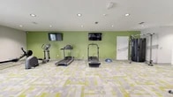 Located at 8737 Baymeadows Rd in Jacksonville, FL, this fitness center offers a comprehensive workout environment across a single, well-organized floor. The space is designed with both functionality and aesthetics in mind, featuring a distinctive geometric-patterned carpet in shades of gray, white, and yellow-green that ties the energetic atmosphere together throughout.

The main exercise area is generously proportioned and equipped with an array of cardio machines, including treadmills, an elliptical, and a stationary bike. A dedicated free weights section features a full rack of dumbbells alongside a multi-functional Training Zone cable and pull-up station. Wall-mounted flat-screen televisions overlook the cardio zone, set against vibrant green accent walls that add a lively, motivating energy to the space. Recessed lighting throughout ensures consistent, bright illumination.

Large glass sliding doors on one side of the main area open to views of an adjacent lounge and outdoor pool area, allowing natural light to flood the space and creating a seamless indoor-outdoor connection.

A separate, quieter exercise room provides space for stretching and balance work, outfitted with stability balls and a wall-mounted Reflect fitness mirror for guided workouts. Natural light enters through a double window, making it ideal for yoga or low-impact training. The facility also includes a private room with a glass-panel door, adding versatility to the overall offering.