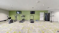 Located at 8737 Baymeadows Rd in Jacksonville, FL, this fitness center offers a comprehensive workout environment across a single, well-organized floor. The space is designed with both functionality and aesthetics in mind, featuring a distinctive geometric-patterned carpet in shades of gray, white, and yellow-green that ties the energetic atmosphere together throughout.

The main exercise area is generously proportioned and equipped with an array of cardio machines, including treadmills, an elliptical, and a stationary bike. A dedicated free weights section features a full rack of dumbbells alongside a multi-functional Training Zone cable and pull-up station. Wall-mounted flat-screen televisions overlook the cardio zone, set against vibrant green accent walls that add a lively, motivating energy to the space. Recessed lighting throughout ensures consistent, bright illumination.

Large glass sliding doors on one side of the main area open to views of an adjacent lounge and outdoor pool area, allowing natural light to flood the space and creating a seamless indoor-outdoor connection.

A separate, quieter exercise room provides space for stretching and balance work, outfitted with stability balls and a wall-mounted Reflect fitness mirror for guided workouts. Natural light enters through a double window, making it ideal for yoga or low-impact training. The facility also includes a private room with a glass-panel door, adding versatility to the overall offering.