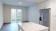 Located at 1001 NE Baker Rd in Stuart, Florida, this well-appointed single-floor residence showcases a clean, contemporary design throughout. The open-concept kitchen is a standout feature, equipped with crisp white shaker cabinetry, stainless steel appliances including a French door refrigerator, dishwasher, and range, complemented by a grey geometric tile backsplash and a generous white-surface island that provides additional prep and dining space. Light wood-style flooring flows seamlessly through the living and bedroom areas, lending warmth to the modern aesthetic. The living area opens directly to a private balcony through a full-height sliding glass door with vertical blinds, inviting natural light and outdoor views. The bedroom is a comfortable, serene space featuring its own large window with blinds and a ceiling fan, as well as access to a walk-in closet with built-in shelving. A second room also features sliding glass door access to the balcony, making it a versatile flex space. The bathroom is tastefully finished with marble-look tile flooring, a soaking tub and shower clad in elongated hexagonal white tile, a warm wood-framed mirror, and a clean white vanity with modern fixtures. A dedicated entryway with a louvered closet completes the thoughtful layout of this bright, move-in-ready residence.