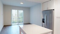 Located at 1001 NE Baker Rd in Stuart, Florida, this well-appointed single-floor residence showcases a clean, contemporary design throughout. The open-concept kitchen is a standout feature, equipped with crisp white shaker cabinetry, stainless steel appliances including a French door refrigerator, dishwasher, and range, complemented by a grey geometric tile backsplash and a generous white-surface island that provides additional prep and dining space. Light wood-style flooring flows seamlessly through the living and bedroom areas, lending warmth to the modern aesthetic. The living area opens directly to a private balcony through a full-height sliding glass door with vertical blinds, inviting natural light and outdoor views. The bedroom is a comfortable, serene space featuring its own large window with blinds and a ceiling fan, as well as access to a walk-in closet with built-in shelving. A second room also features sliding glass door access to the balcony, making it a versatile flex space. The bathroom is tastefully finished with marble-look tile flooring, a soaking tub and shower clad in elongated hexagonal white tile, a warm wood-framed mirror, and a clean white vanity with modern fixtures. A dedicated entryway with a louvered closet completes the thoughtful layout of this bright, move-in-ready residence.