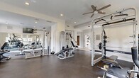 This fitness center at 1755 S Beeler St in Denver, Colorado, is a comprehensive, single-floor facility designed to support a full range of workout routines. The space is divided into distinct functional zones, providing an organized and efficient layout for users of all fitness levels.

The strength training area is generously sized and features a power rack with weighted barbells, multiple cable and pulley machines, adjustable benches, and a full dumbbell rack, offering extensive options for resistance training. Large wall-mounted mirrors line one section of the space, enhancing the sense of openness while allowing users to monitor their form.

A dedicated cardio zone houses an impressive selection of equipment, including stair climbers, elliptical trainers, a stationary upright bike, a recumbent bike, and a rowing machine. This area benefits from multiple large windows fitted with roller shades, flooding the space with natural light and providing views of the surrounding greenery.

Throughout the facility, durable rubber flooring provides traction and support, while recessed lighting and ceiling fans maintain a comfortable environment. Structural columns divide the space into approachable sections without disrupting the overall flow. A tiled entryway corridor connects the main workout areas, and a direct exterior door provides convenient access. The clean, white walls and modern finishes give the space a bright, professional ambiance well-suited for daily use.