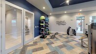 The Health Hub at 6255 Fernwood Dr. in Kannapolis, North Carolina is a thoughtfully designed, single-floor fitness center that offers a comprehensive workout experience. The space is finished with a distinctive patterned carpet in neutral tones of beige, gray, and blue, lending the facility a clean and contemporary aesthetic throughout.

The cardio zone features a row of treadmills, an elliptical, and a stair climber, all positioned beneath a bold navy-blue tray ceiling and accompanied by wall-mounted flat-screen televisions. Multiple windows along the perimeter fill the space with natural light. A prominently displayed "Health Hub" logo anchors this section of the room.

The strength training area is equipped with a Smith machine and adjustable bench, providing a dedicated space for resistance and weightlifting workouts. A built-in countertop with cabinetry and a large mirror along one wall adds both function and visual depth to this zone.

A functional training corner showcases a tiered rack stocked with medicine balls, kettlebells, resistance tools, and stability balls, alongside a dumbbell rack and a rowing machine. Motivational fitness-themed typography adorns the wall, reinforcing the space's energetic atmosphere.

Recessed lighting throughout ensures consistent illumination, while a ceiling fan supports air circulation. A glass-panel door provides easy access, and the overall layout supports a smooth, efficient flow between all workout zones.