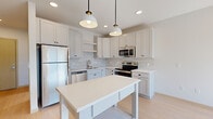 This single-floor residence is designed with a clean, contemporary aesthetic, featuring light hardwood flooring and a bright, neutral palette throughout. The open-concept kitchen serves as the heart of the space, outfitted with white shaker-style cabinetry, a chevron-patterned marble tile backsplash, quartz countertops, and a full suite of stainless steel appliances including a refrigerator, electric range, microwave, and dishwasher. A freestanding white kitchen island with a lower shelf provides additional workspace and storage, illuminated by a pair of stylish pendant lights. The living area is bathed in natural light from large sliding glass doors that open to a private balcony, offering outdoor views and ventilation supplemented by a wall-mounted split AC unit. The bedroom features a tall window that draws in abundant sunlight. The bathroom includes a single-sink vanity with white cabinetry and quartz countertop, a walk-in shower, and built-in shelving. Throughout the unit, consistent hardwood flooring, clean white walls, and modern fixtures contribute to a cohesive, move-in-ready atmosphere.