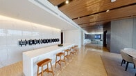 This refined single-floor space is designed with a seamless blend of warm wood accents, marble surfaces, and polished stone flooring that establishes a consistently luxurious aesthetic throughout. The central lounge area features slatted wood ceilings with recessed lighting, a built-in media wall flanked by curated shelving, and generous seating arrangements. Large floor-to-ceiling sliding glass doors open directly to an outdoor terrace, filling the interior with natural light and providing expansive outdoor views.

A sleek marble-topped bar runs along one wall, lined with leather-and-wood barstools and a backlit wine display, creating an elegant entertaining focal point. Adjacent to this, an open dining area with a round table and upholstered chairs also connects to the terrace through full-height glazing. A dedicated lounge zone with a wall-mounted screen and built-in banquette seating offers additional gathering space.

The private meeting room features a substantial wooden conference table, roller blinds, a suspended linear pendant, and views to the outdoors. A reception nook with curated artwork and lounge chairs provides a polished transitional area. The catering kitchen offers dual-sided countertops, high-gloss upper cabinetry, under-cabinet lighting, and a marble backsplash for full-service functionality.