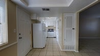 This single-floor space presents a practical and well-organized layout centered around a functional kitchen. The kitchen is outfitted with crisp white cabinetry that extends to the upper walls, providing ample storage throughout. White appliances, including a full-size refrigerator and a gas range with an overhead exhaust hood, are neatly integrated into the space, contributing to a cohesive and tidy aesthetic. Light-colored ceramic tile flooring runs throughout, lending a bright and easy-to-maintain surface underfoot. A tray ceiling detail adds a subtle architectural element, giving the entry area a slightly elevated sense of dimension. Ventilation is well-addressed with ceiling-mounted HVAC registers visible throughout. A louvered door adds a classic touch and provides additional storage or utility access. The neutral wall tones in soft beige complement the white cabinetry and appliances, creating a clean, understated palette. The open sightline from the entry through to the kitchen enhances the sense of depth and flow, making the space feel connected and functional. Overall, the layout prioritizes efficiency and practicality with a straightforward, no-frills design.