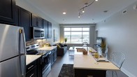 This stylish single-floor residence showcases a thoughtfully designed open-concept layout that seamlessly connects the kitchen, dining, and living areas into one cohesive living space. The kitchen is a standout feature, outfitted with dark, bold cabinetry contrasted by crisp white countertops, a classic tile backsplash, and a full suite of stainless steel appliances including a built-in microwave, electric range, and dishwasher. A sleek kitchen island with pendant-style track lighting overhead serves as both a functional workspace and a casual dining destination. The living area is anchored by a large picture window that floods the space with natural light and frames sweeping views of the surrounding cityscape and landscape beyond. Neutral gray walls and polished concrete-style flooring lend a clean, contemporary aesthetic throughout, while recessed lighting adds warmth and depth. The open flow between spaces makes the layout feel spacious and versatile, ideal for both everyday living and entertaining. Overall, the unit strikes a refined urban balance between modern design and practical comfort.
