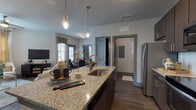 This single-floor residence presents a cohesive, contemporary design throughout its open-concept layout. The kitchen serves as a central focal point, featuring rich dark cabinetry, granite countertops, a large island with bar seating, stainless steel appliances including a refrigerator, electric range, and built-in microwave, plus a white subway tile backsplash. Pendant lighting with an industrial-style finish illuminates the island, while wood-look flooring extends throughout the main living areas, creating visual continuity.

The dining area adjoins the kitchen and offers direct sightlines into the living room, enhancing the open flow of the space. The living room is bright and welcoming, benefiting from multiple windows with blinds and drape treatments, and features a ceiling fan overhead. A separate sitting nook provides additional flexible living space with natural light from large windows.

The bedrooms are carpeted for comfort, each accommodating generous furniture and benefiting from natural light. One bedroom includes a walk-in closet. The bathroom is well-appointed with granite vanity surfaces, dark cabinetry, wood-look flooring, and a full tub-shower combination with white tile surround. Neutral gray tones and dark trim details carry a consistent, modern aesthetic throughout the entire residence.